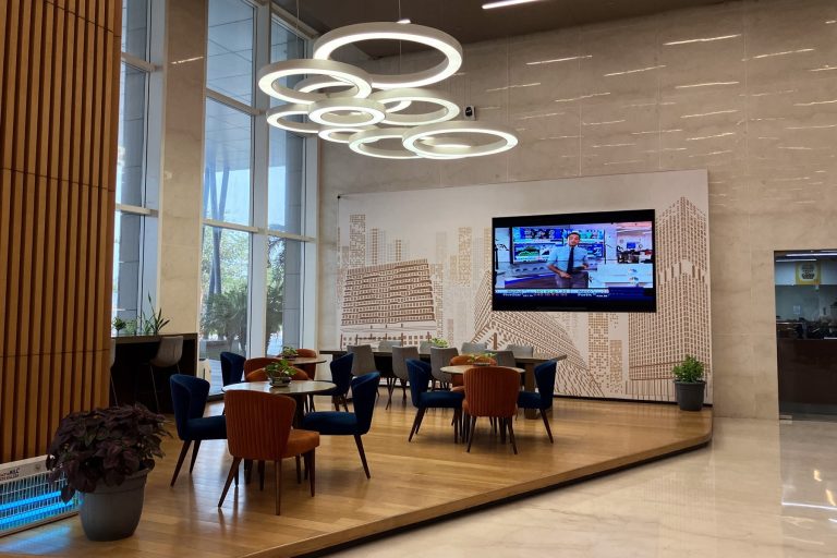 A lobby area with chairs arranged around several working height tables. A large widescreen television is fixed to a wall featuring architectural artwork.