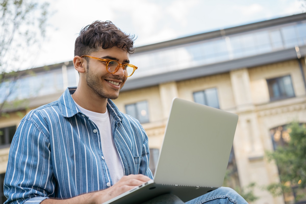 A smiling student works outside with a laptop on their knee